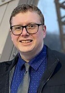 Sam Monson smiles wearing glasses, a blue button-up, and a black jacket.