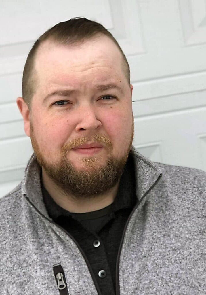 Sean Hamilton smiles wearing a gray quarter zip in front of a garage door as the background.