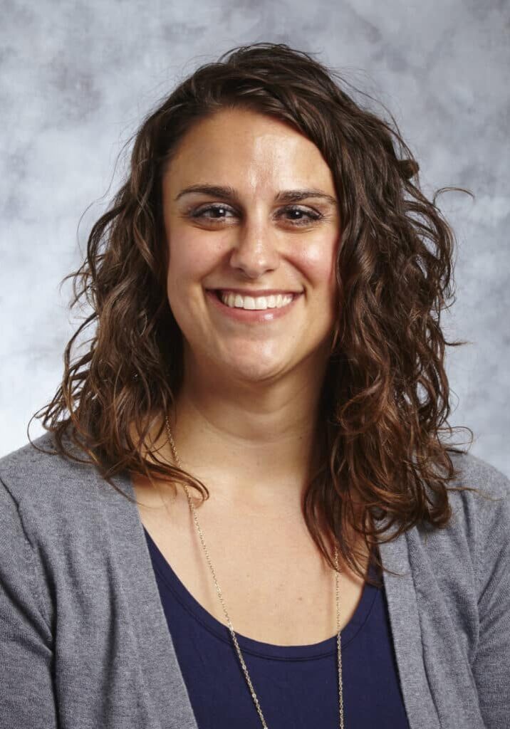 Stacy Gross smiles in front of a gray portrait background wearing a gray sweater over a navy blue top.