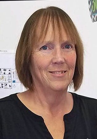 Sue Featherly smiles wearing a black blouse standing in front of a white wall.