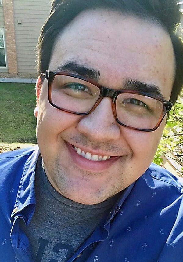 Tom Brinker smiles wearing a blue button up and brown glasses.