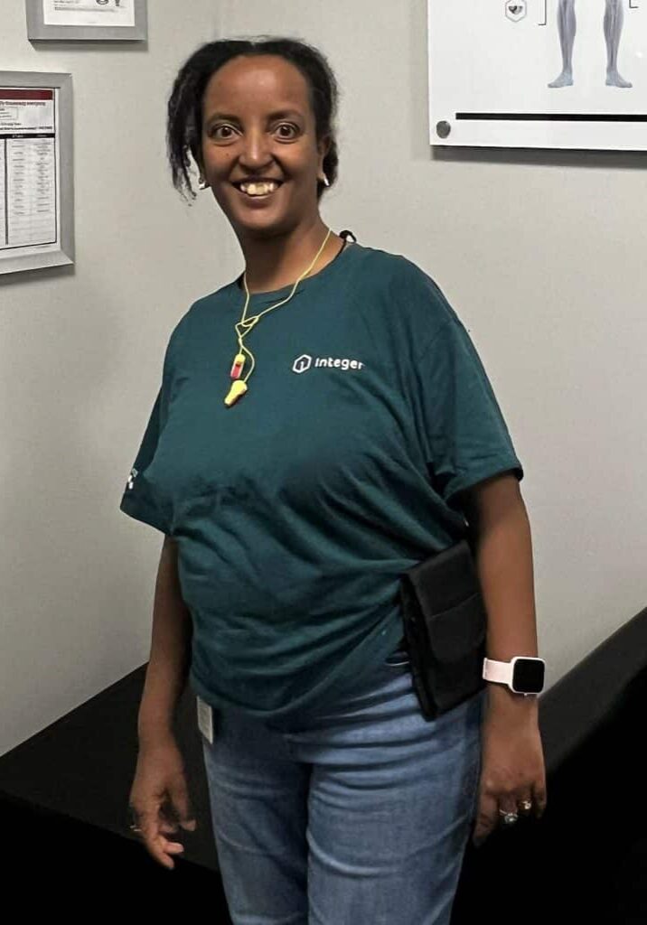 Tsega smiles in front of a gray wall wearing a dark teal shirt.