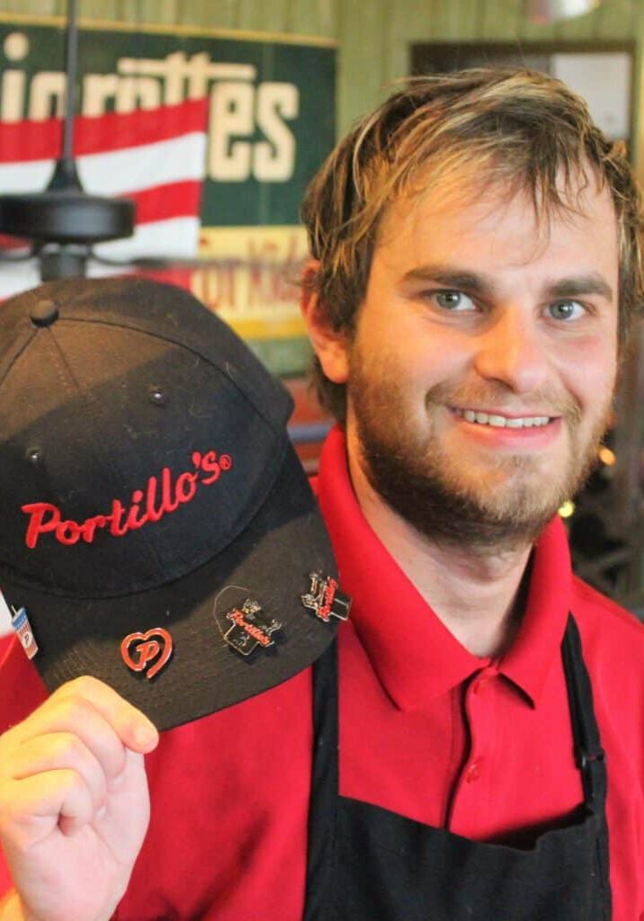 Tyler Wollersheim smiles at his workplace holding his Portillo's work hat and red polo shirt.