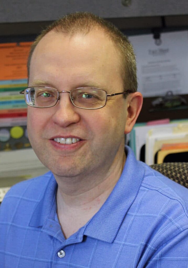 Vince Mans sits in his desk smiling wearing glasses and a blue shirt.