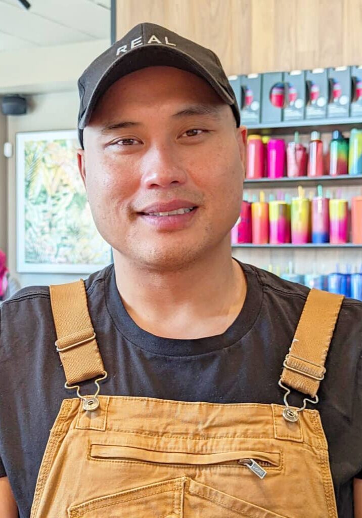 Hue Yang smiles wearing tan overalls in front of a wall of colorful Starbucks cups.