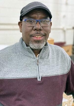 Ali Al-Sadi smiles wearing glasses, a hat, and a gray/maroon quarter-zip sweater.