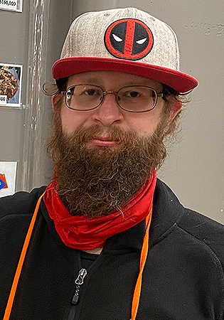 Rob Flood smiles in front of a gray background wearing glasses and a baseball hat.