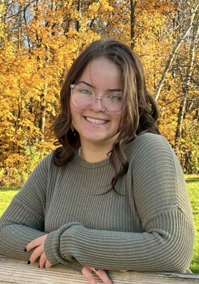 Camryn Warren smiles for the camera in front of yellow autumn foliage.